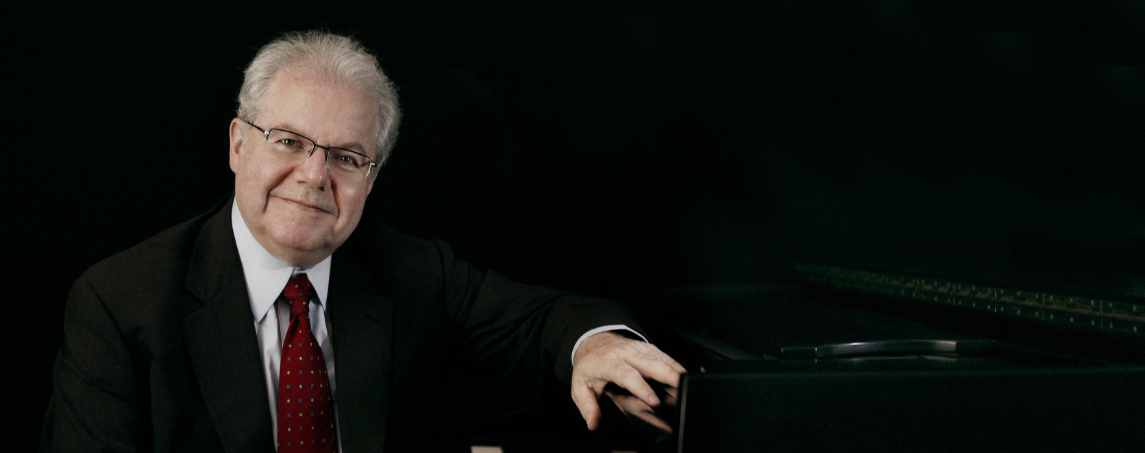 An older man with short gray hair and glasses sits beside a piano against a dark background. He wears a dark suit, white shirt, and red tie, and rests one hand casually on the piano. He looks toward the camera with a calm, gentle expression.